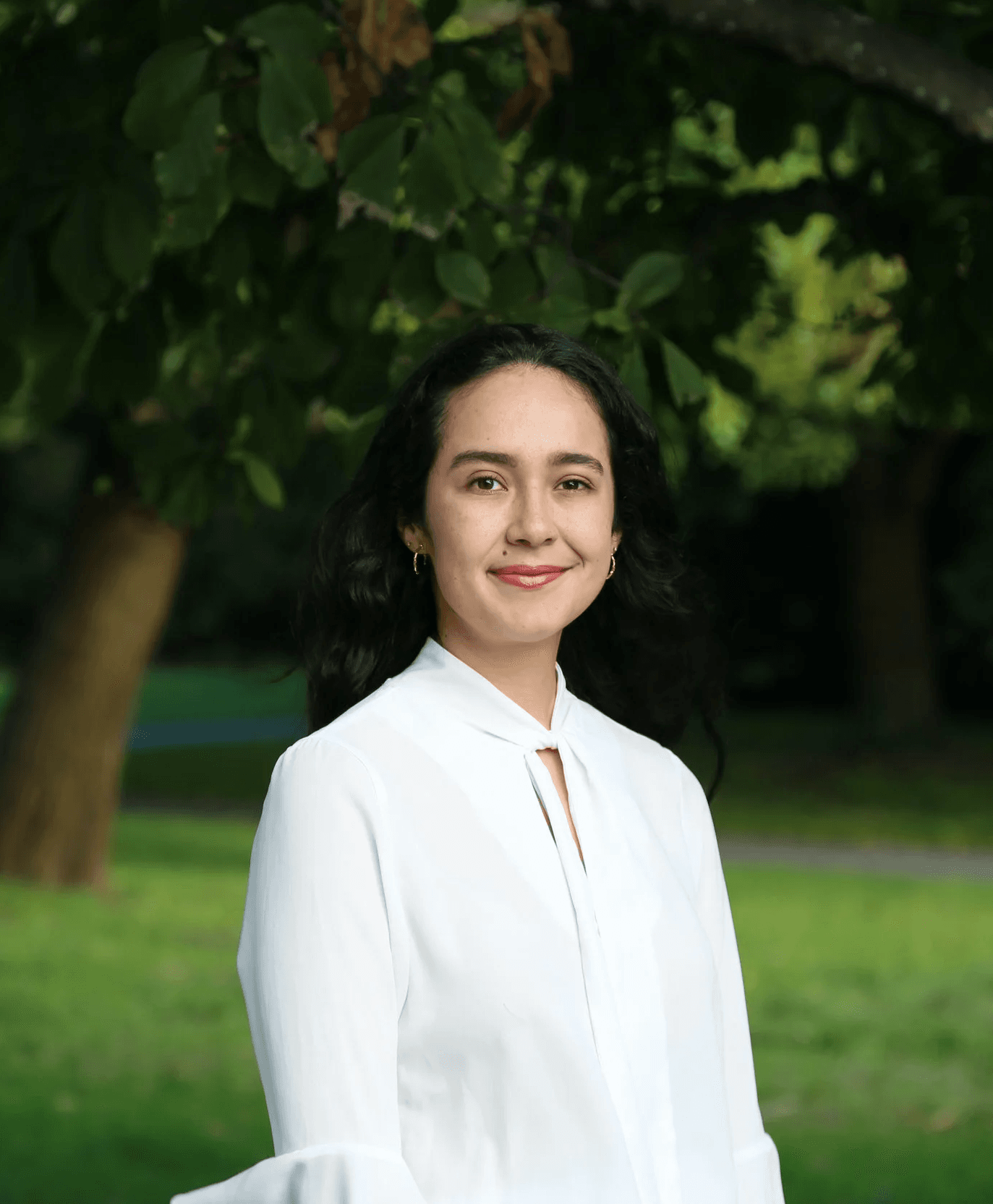 A young woman with dark, wavy hair and earrings, wearing a white blouse, stands outdoors with trees and grass in the background.